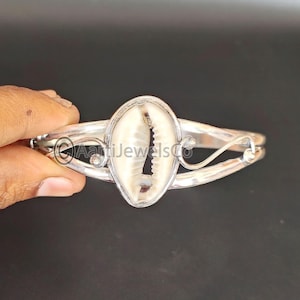 Może przedstawiać: Srebrna bransoletka z zawieszką w kształcie białej muszli kauri. Bransoletka ma delikatny, zakrzywiony wzór.