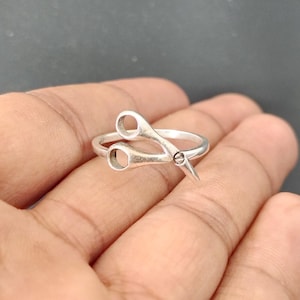 以下が含まれることがあります： ハサミの形をしたシルバーカラーのリング。リングは、開いたハンドルと尖った刃を備えたハサミの詳細なデザインが特徴です。リングは指に着用され、複雑な職人技とユニークなスタイルを披露しています。