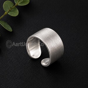 Può includere: Un anello color argento con una texture in metallo spazzolato. L'anello ha una fascia larga che si curva attorno al dito, con un design aperto. L'anello è esposto su una superficie nera.