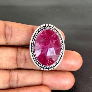 May include: A silver ring featuring a large, faceted, oval-shaped ruby gemstone. The ruby is a deep, vibrant red color. The ring has a decorative silver border with a rope-like design.