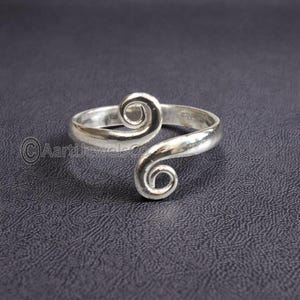 Può includere: Un anello per il dito del piede color argento con un design a spirale. L'anello ha una fascia curva che avvolge il dito del piede, con una spirale nella parte superiore e un'altra all'estremità della fascia. L'anello è su uno sfondo scuro.