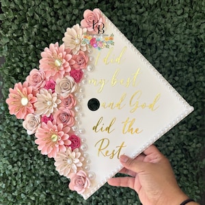 Könnte beinhalten: Eine weiße Abschlusskappe, verziert mit rosa und cremefarbenen Papierblumen, Perlen und goldfarbenen Buchstaben mit der Aufschrift "I did my best and God did the Rest". Die Kappe hat ein schwarzes Loch in der Mitte.