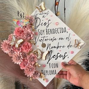 Op de afbeelding: Een versierde afstudeerhoed met roze papieren bloemen, gouden accenten en vlinderversieringen. De hoed bevat de tekst "Gracias a Dios, Soy Bendecida, Victoriosa en el Nombre de Jesus, Y con Diploma."