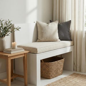 May include: A white bench with a striped cushion and two pillows in beige and gray. A woven basket sits below the bench. A small wooden side table holds a candle, books, and a vase with greenery.