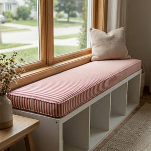 May include: A white rectangular storage bench with four open cubby compartments, topped with a long, firm cushion featuring a red and white vertical striped pattern. A single beige textured throw pillow rests on the right side of the bench.