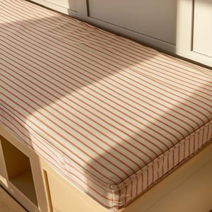 May include: A rectangular bench cushion with a cream-colored background and evenly spaced red stripes. The cushion is placed on a light-colored bench with storage compartments. The fabric has a classic, farmhouse style.