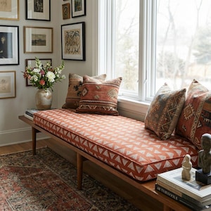 May include: A wooden bench with a rust-colored cushion featuring a white triangle pattern. Decorative pillows with patterned designs in shades of brown and orange rest on the bench. A vase of flowers and framed artwork are visible in the background.