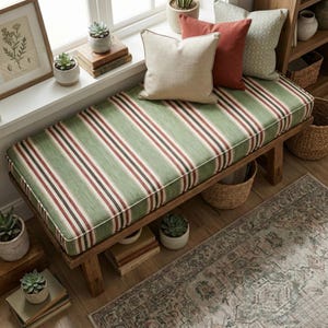 Op de afbeelding: Een houten bank met een groen en wit gestreept kussen, geaccentueerd met rode en zwarte strepen. Drie kussens in beige, roest en groen met patronen liggen op de bank. De bank is omgeven door planten, boeken en een vloerkleed.