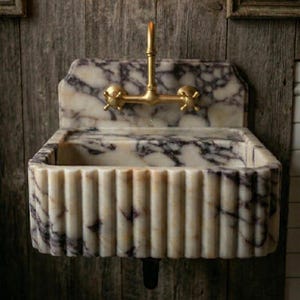 May include: A marble sink with a fluted front and a gold-coloured tap. The sink has a rectangular basin and a backsplash. The marble has a white and purple veined pattern. The tap has two cross-shaped handles.