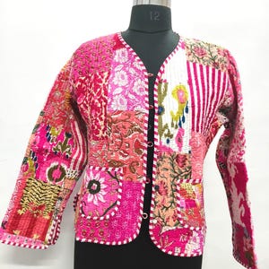 May include: A patchwork jacket in shades of pink, red, and white, with floral and striped patterns. The jacket has a V-neck, long sleeves, and two front pockets. The edges are trimmed with a white and red dotted border. The jacket is displayed on a black mannequin.