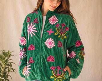 May include: A green velvet zip-up jacket featuring vibrant pink and yellow floral embroidery across the front and sleeves. The jacket has a high collar, long sleeves with elastic cuffs, and two side pockets, worn over a white shirt and blue jeans.
