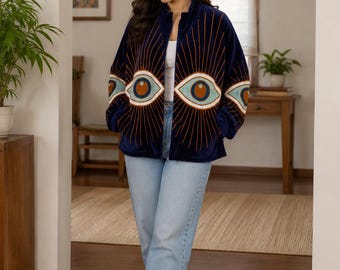 Puede incluir: Chaqueta de terciopelo azul marino con cuello alto y cremallera. La chaqueta presenta un diseño de ojos repetido en tonos marrón, naranja y blanco. La modelo lleva vaqueros azul claro y zapatillas blancas.