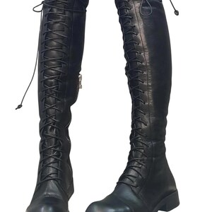 May include: A pair of black leather knee-high boots. The boots feature a lace-up design that extends from the foot to the top of the knee. A zipper is visible on the side of one boot. The boots have a flat sole.