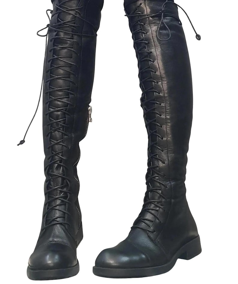 May include: A pair of black leather knee-high boots. The boots feature a lace-up design that extends from the foot to the top of the knee. A zipper is visible on the side of one boot. The boots have a flat sole.