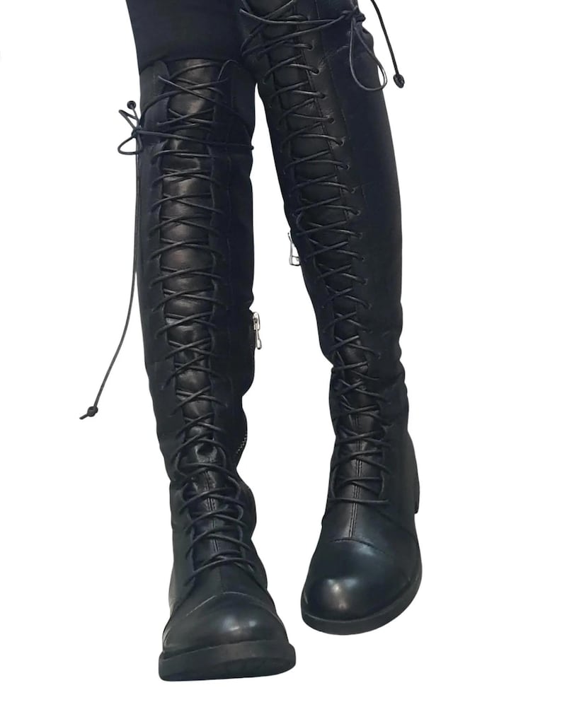 May include: Black, knee-high boots with a lace-up design. The boots have a rounded toe and a zipper on the side. The laces extend up the front of the boots. The boots are made of a smooth, leather-like material.