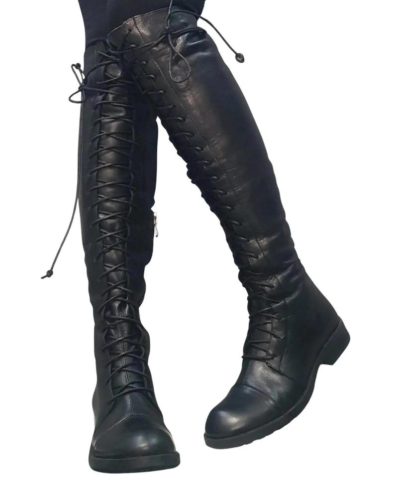 May include: Black leather knee-high boots with lace-up detailing. The boots have a rounded toe and a low heel. The laces extend up the front of the boots. The boots are made of a smooth, matte leather.
