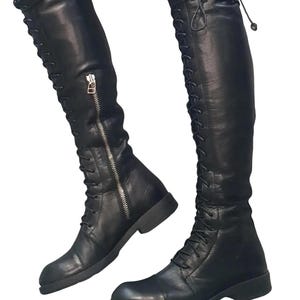 May include: Black leather over-the-knee boots with a lace-up design and a side zipper. The boots have a flat sole and a sleek, modern style. The boots are made of a smooth, shiny leather material.