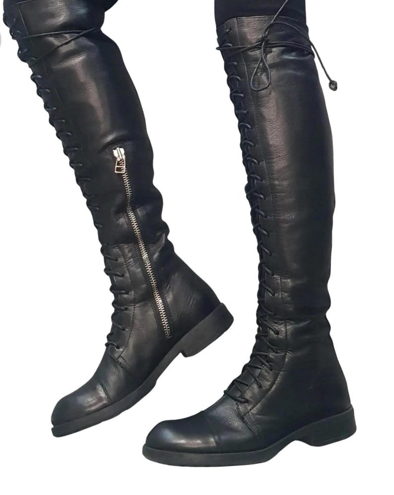 May include: Black leather over-the-knee boots with a lace-up design and a side zipper. The boots have a flat sole and a sleek, modern style. The boots are made of a smooth, shiny leather material.