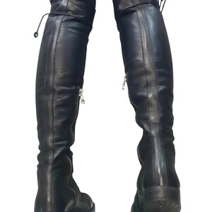 May include: Black leather over-the-knee boots with silver zippers and laces at the top. The boots have a flat sole and are designed to be worn over the knee. The boots are made of a smooth, shiny leather material.