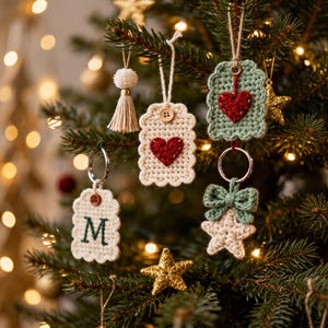 Puede incluir: Una colección de adornos de ganchillo colgados en un árbol, que incluye una etiqueta crema con un corazón rojo y un botón de madera, una etiqueta verde salvia con un corazón rojo, una etiqueta crema con la letra M, una estrella crema y una borla.