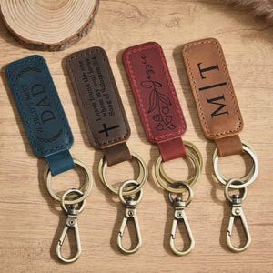 May include: Four leather keyrings with metal fixings. One is blue with "WORLD'S BEST DAD" text, another brown with a cross and text, a red one with "Jessie" and floral design, and a tan one with "M|T".