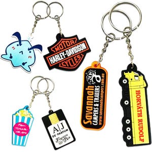 Könnte beinhalten: Eine Sammlung von Schlüsselanhängern mit verschiedenen Designs. Enthält ein Harley-Davidson-Logo, eine Cartoonfigur, einen Popcorn-Eimer, eine Parfümflasche, ein Camper-Logo und einen gelben LKW. Jeder Schlüsselanhänger hat einen Metallring und eine Kette.