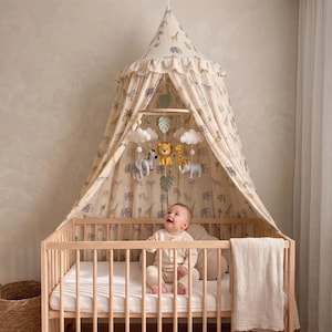 May include: A beige fabric canopy with a safari animal print featuring lions, elephants, and giraffes hangs over a wooden cot. A mobile with plush animal figures and white clouds is suspended inside the canopy, which has a ruffled top edge.