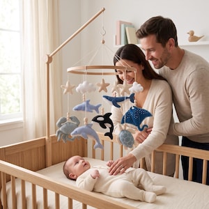 May include: A wooden baby mobile featuring felt sea creatures including a whale, dolphin, orca, crab, sea turtle, and stingray, interspersed with felt stars and clouds, suspended from a circular wooden frame on a wooden arm stand.