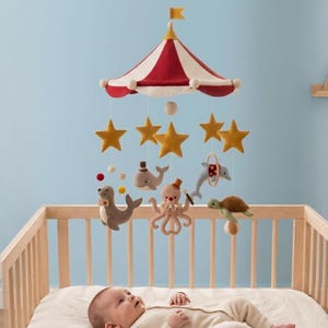 May include: A hanging nursery mobile featuring a red and cream striped circus tent canopy with a small flag on top. Suspended from the canopy are yellow felt stars and plush sea creatures including a seal, whale, octopus, dolphin, and turtle.
