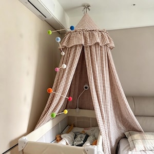 May include: A beige and white gingham fabric canopy with a ruffled top edge, suspended over a crib. A hanging mobile with thin wooden rods and colorful felt spheres in blue, green, white, pink, orange, and yellow is positioned to the left side.