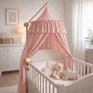 May include: A white wooden cot featuring a pink and white gingham fabric canopy suspended from a wooden arm. The canopy has a ruffled top edge. Inside the cot, there is a light brown plush teddy bear and a star-shaped pillow on a white mattress.
