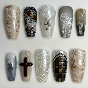 May include: A collection of ten artificial nails with various designs. The nails feature intricate patterns, including stained glass, crosses, floral motifs, and abstract art. Colors include gold, white, brown, gray, and black. The nails are oval-shaped and have a glossy finish.