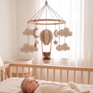 May include: A hanging nursery mobile featuring a circular wooden frame with suspended beige felt clouds, crescent moons, stars, and a central hot air balloon carrying a small brown teddy bear. The elements are attached with thin brown cords.