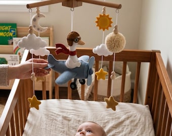 May include: A nursery mobile featuring a felt goose pilot wearing brown goggles and a red scarf, seated in a blue felt airplane. The mobile includes hanging felt elements: a gray moon, white clouds, a yellow sun, yellow stars, and a beige pom-pom.