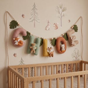 May include: A felt letter garland spelling CHLOE in muted tones of pink, sage green, yellow, and tan. Each letter features a small woodland animal figure, including a fox, bear, deer, owl, and rabbit, interspersed with green felt leaves and beads.