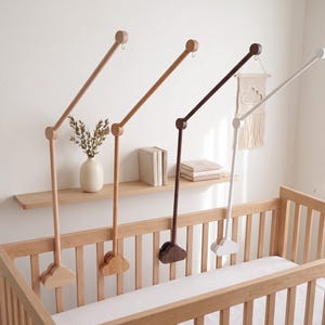 May include: Four wooden cot mobile arms in light wood, medium wood, dark wood, and white finishes. Each arm features a cloud-shaped base that attaches to the cot rail, a jointed middle section, and a long angled top arm with a metal hook at the end.