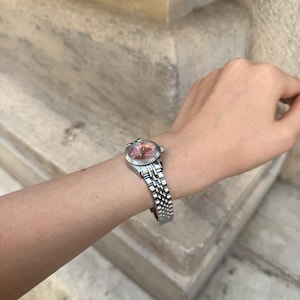 Puede incluir: Reloj de pulsera plateado con esfera rosa y correa plateada. El reloj tiene una esfera redonda y una correa texturizada. El reloj est&aacute; en una mu&ntilde;eca.