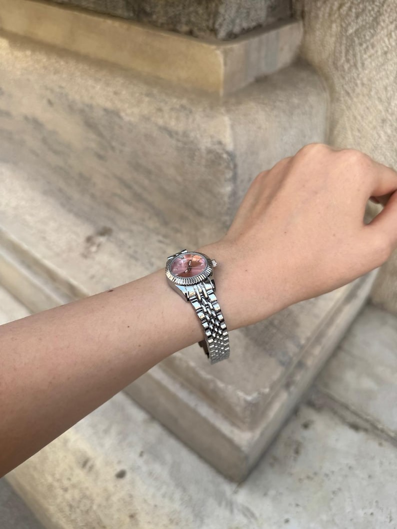 Puede incluir: Reloj de pulsera plateado con esfera rosa y correa plateada. El reloj tiene una esfera redonda y una correa texturizada. El reloj est&aacute; en una mu&ntilde;eca.