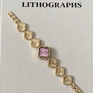 Puede incluir: Un reloj de pulsera dorado con eslabones en forma de trébol y una esfera rosa. El reloj está sobre un libro titulado "PICASSO LITHOGRAPHS".