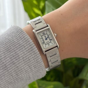 May include: A rectangular silver quartz watch with a white face and black Roman numerals. The watch has a silver strap and is worn on the wrist. The watch face reads "Quartz".