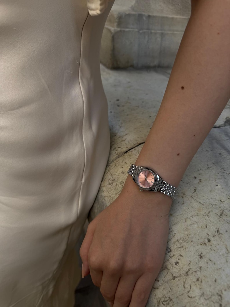 Puede incluir: Un reloj de pulsera plateado con esfera rosa se lleva en la mu&ntilde;eca. El reloj tiene un bisel texturizado y una correa de metal con eslabones. El fondo muestra un vestido blanco y una superficie de piedra.
