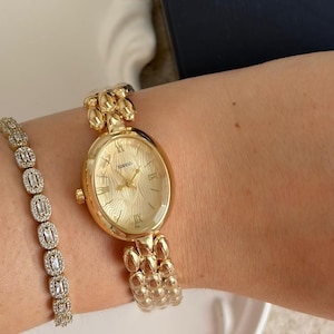 May include: A gold-toned wristwatch with an oval face and Roman numeral hour markers. The watch is paired with a matching gold bracelet with rectangular, clear stones. The watch and bracelet are on a wrist.