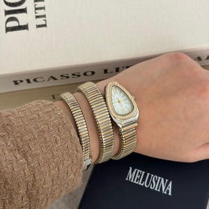 May include: A gold-toned watch with a triangular face and a white dial, adorned with small crystals. The watch is paired with two matching gold bracelets. The watch and bracelets are on a wrist, with a box that says "MELUSINA" in the background.