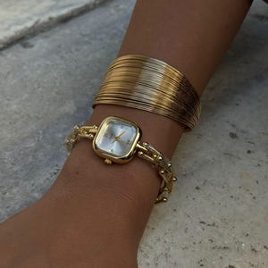Puede incluir: Un reloj dorado con esfera cuadrada y delicada pulsera de cadena se lleva en la muñeca, combinado con una pulsera ancha dorada. La esfera del reloj es de color claro y las pulseras son brillantes.