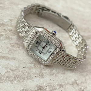 Puede incluir: Reloj de pulsera plateado con esfera cuadrada y brazalete de eslabones. La esfera es blanca con números romanos negros y un logotipo de marca. El bisel está incrustado con pequeños cristales transparentes. Una pequeña gema azul adorna la corona.