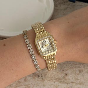 May include: A gold-toned watch and bracelet set. The watch has a rectangular face with a white dial, Roman numerals, and a diamond-studded bezel. The bracelet features rectangular, clear stones. The watch band and bracelet are linked.