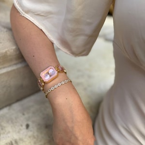 Puede incluir: Un reloj dorado con una esfera rectangular rosa y detalles rosas a juego. También se ve una delicada pulsera dorada con pequeños adornos redondos. El reloj y la pulsera se llevan en la muñeca de una persona, junto con un anillo.