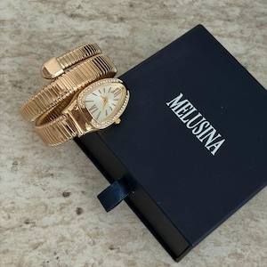 May include: A gold-toned wristwatch with a snake-like design, featuring a white watch face with gold hands and Roman numerals. The watch is next to a navy blue box with the word "MELUSINA" printed on it.