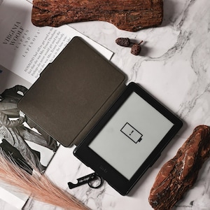 Pu&ograve; includere: Un e-reader nero con una custodia marrone &egrave; sdraiato su una superficie di marmo bianco. Lo schermo dell'e-reader mostra un'icona di batteria scarica. L'e-reader &egrave; circondato da elementi naturali, tra cui un pezzo di legno di deriva, pigne e uno stelo d'erba secca.