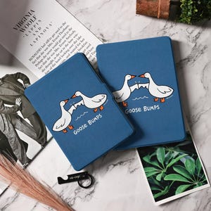May include: Two blue rectangular cases with a cartoon goose design and the words "Goose Bumps" in white. The geese are white with orange feet and beaks. The cases are on a marble surface.